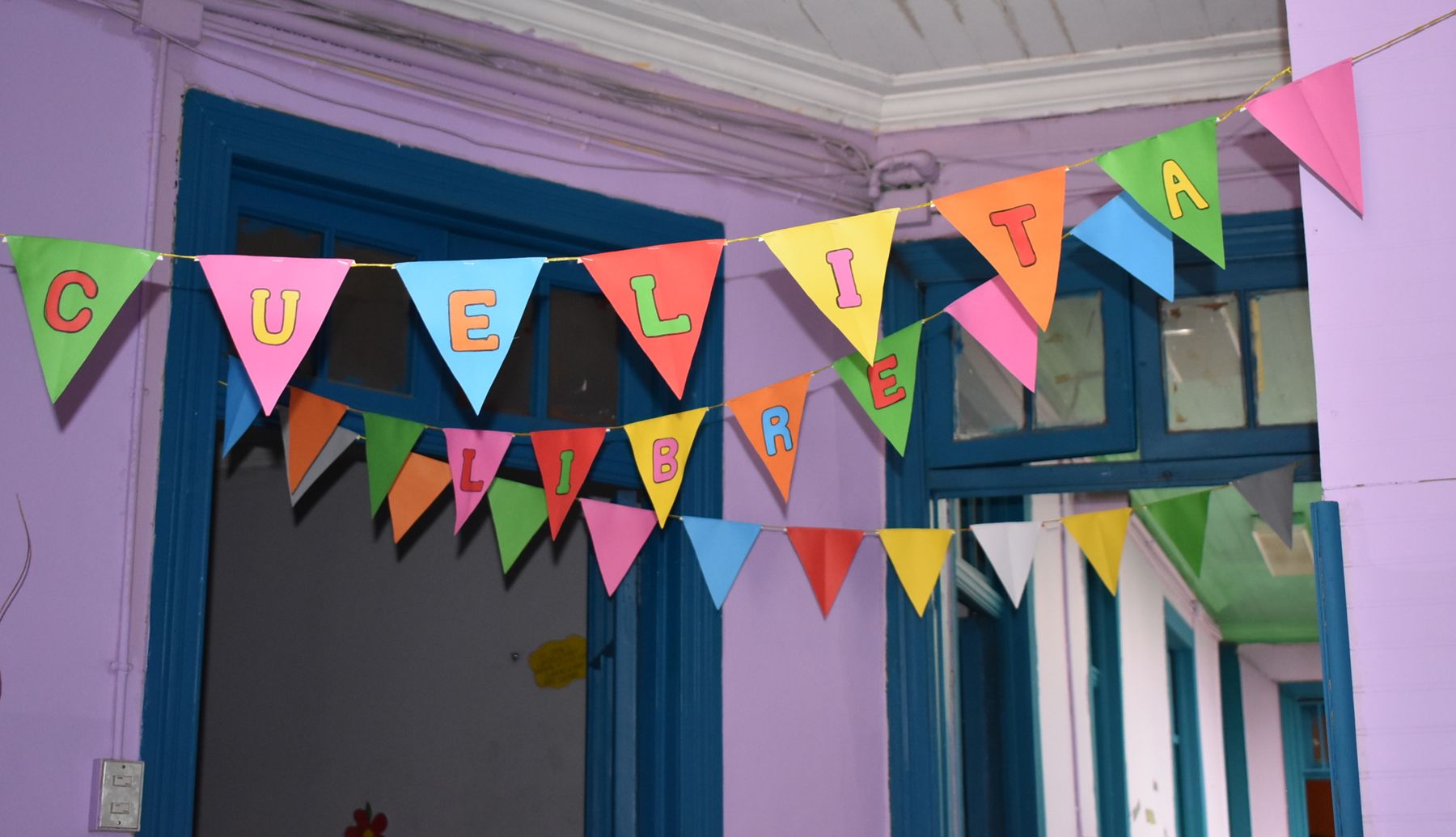 Banderines de papel de colores colgados frente a la puerta azul de la sede, en una pared lila